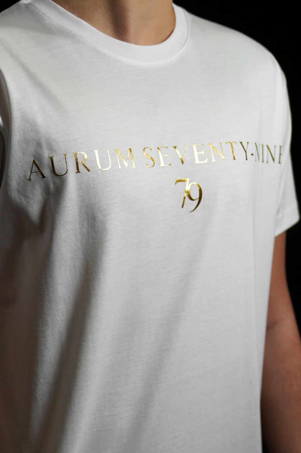 Aurum Seventy-nine: Vista leggermente laterale di un modello che indossa la T-shirt Round Neck Aurum Classic 79 White, con focus sul logo dorato al centro del petto e il girocollo a costina.