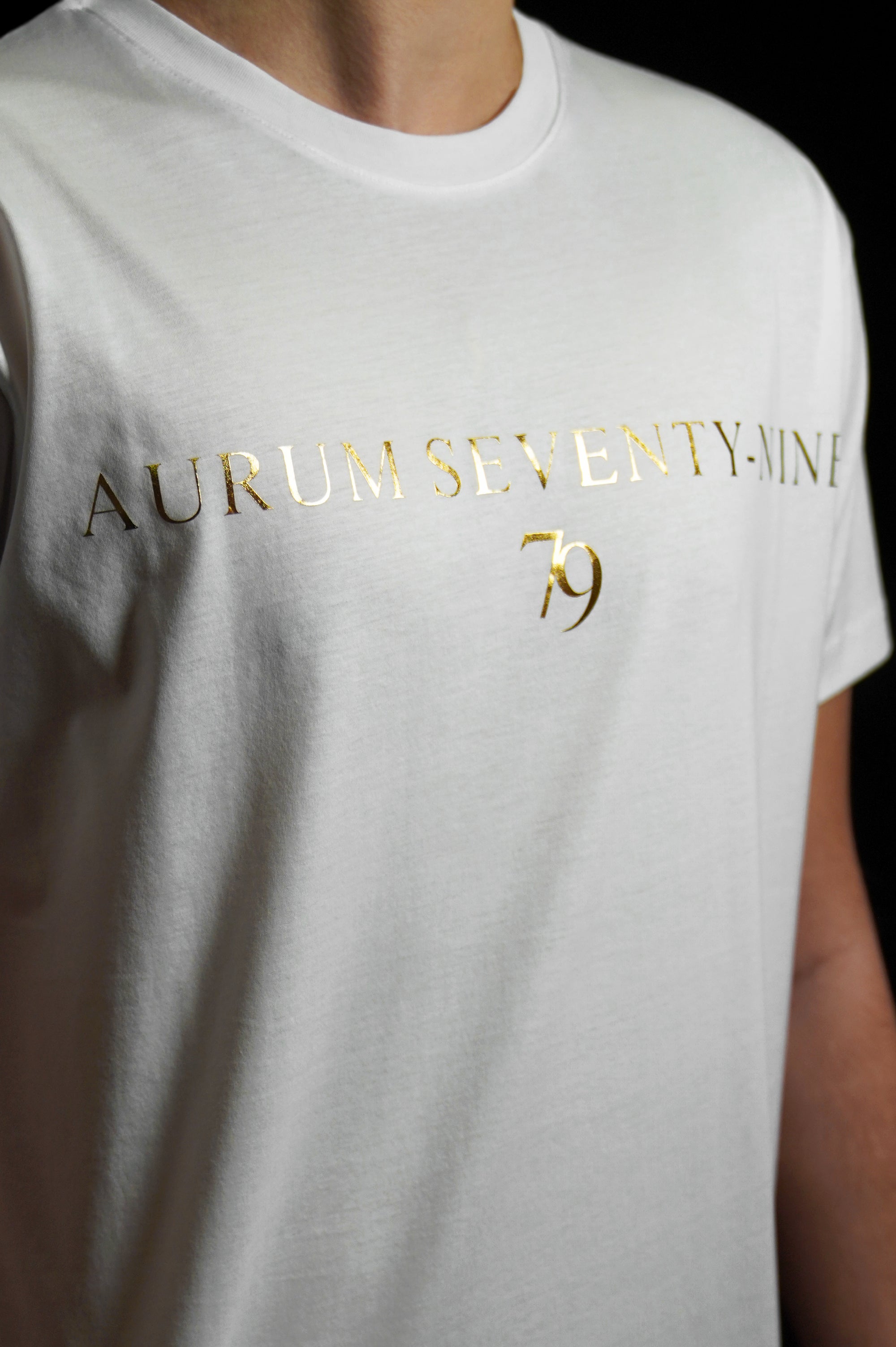 Aurum Seventy-nine: Vista leggermente laterale di un modello che indossa la T-shirt Round Neck Aurum Classic 79 White, con focus sul logo dorato al centro del petto e il girocollo a costina.