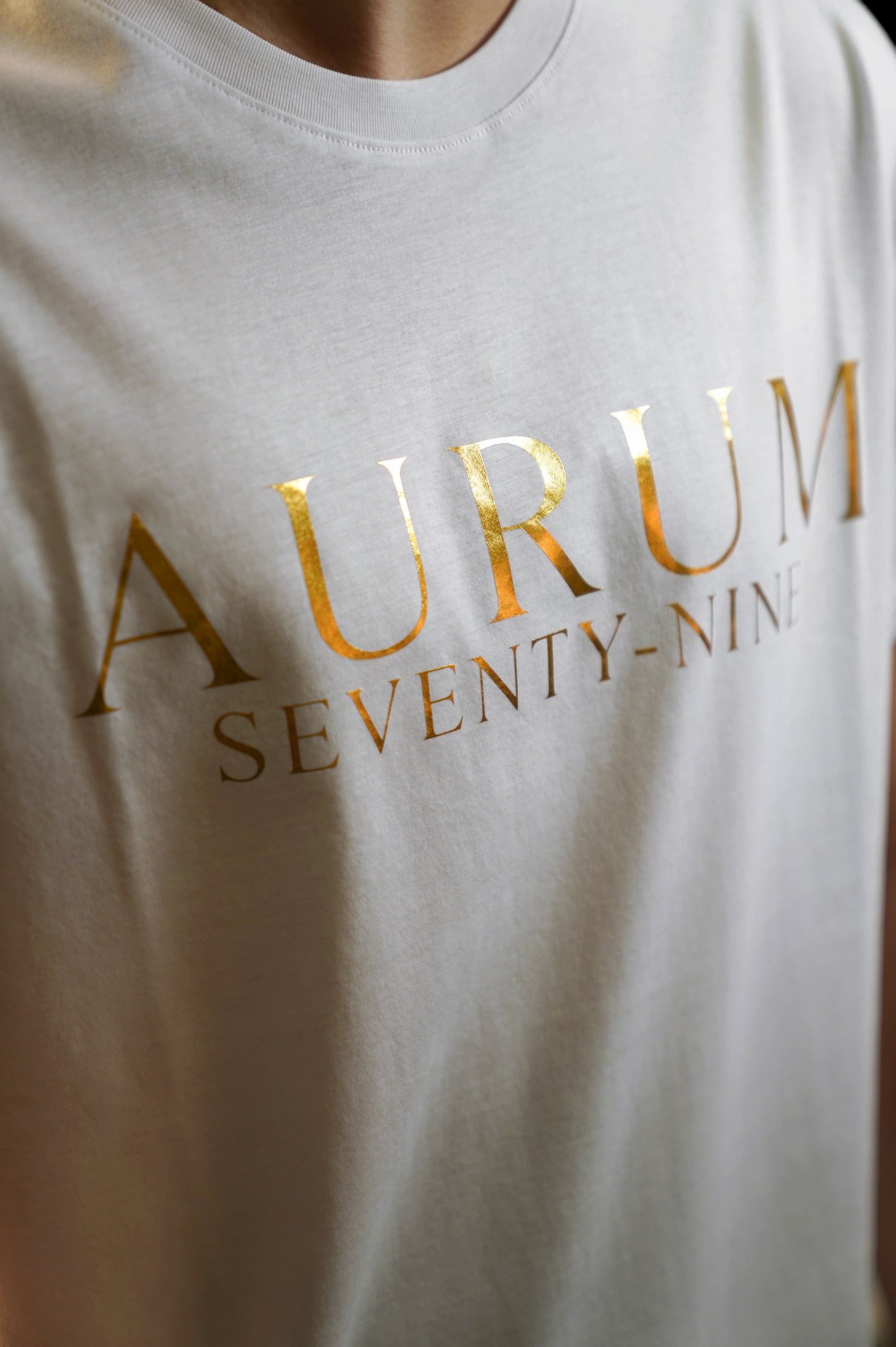 Aurum Seventy-nine: Vista leggermente laterale di un modello che indossa la T-shirt Round Neck Aurum Classic White, con focus sul logo dorato al centro del petto e il girocollo a costina.