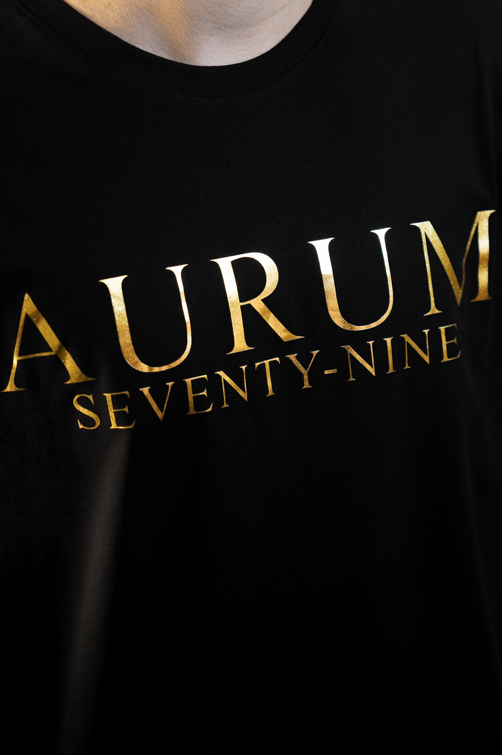Aurum Seventy-nine: Vista leggermente laterale di un modello che indossa la T-shirt Round Neck Aurum Classic Black, con focus sul logo dorato al centro del petto e il girocollo a costina.