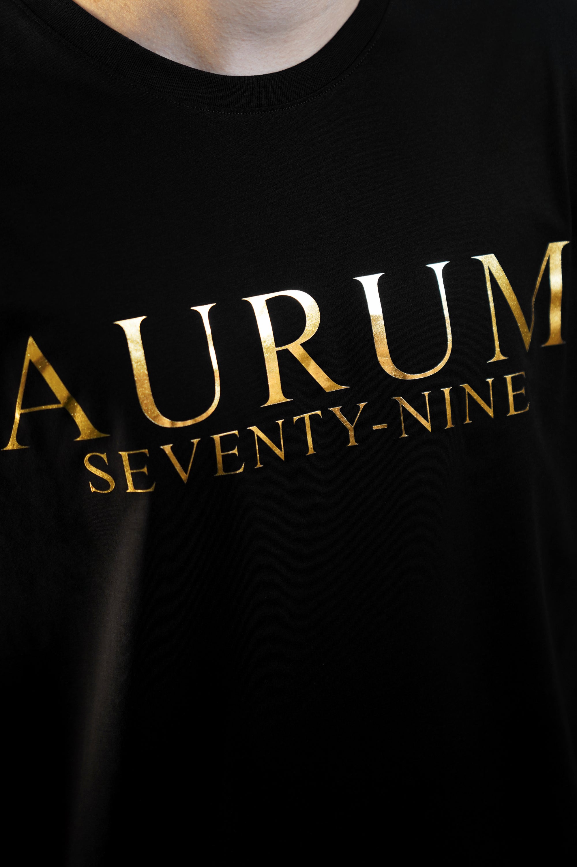 Aurum Seventy-nine: Vista leggermente laterale di un modello che indossa la T-shirt Round Neck Aurum Classic Black, con focus sul logo dorato al centro del petto e il girocollo a costina.