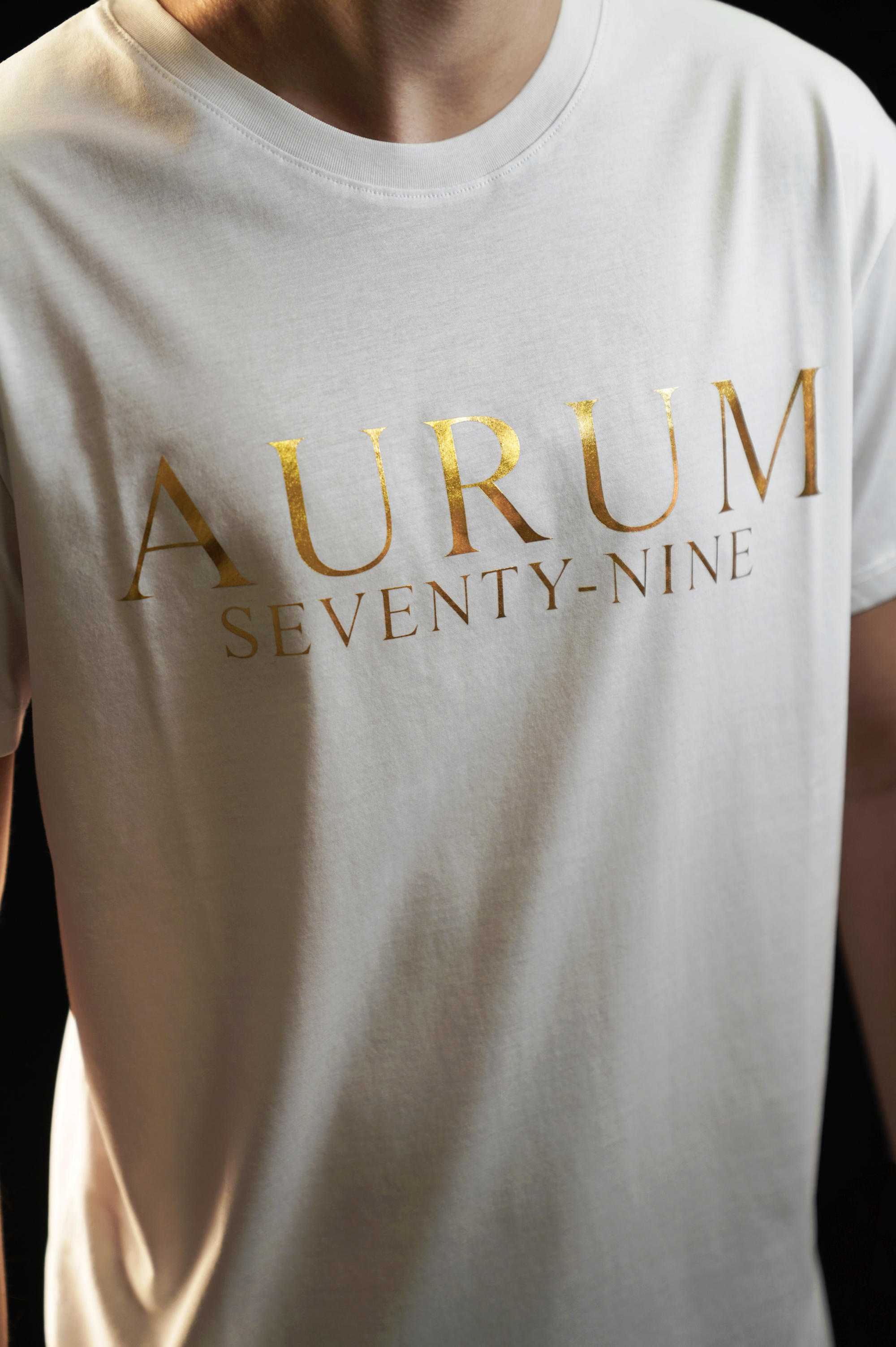 Aurum Seventy-nine: Vista leggermente laterale di un modello che indossa la T-shirt Round Neck Aurum Classic White, con focus sul logo dorato al centro del petto e il girocollo a costina.