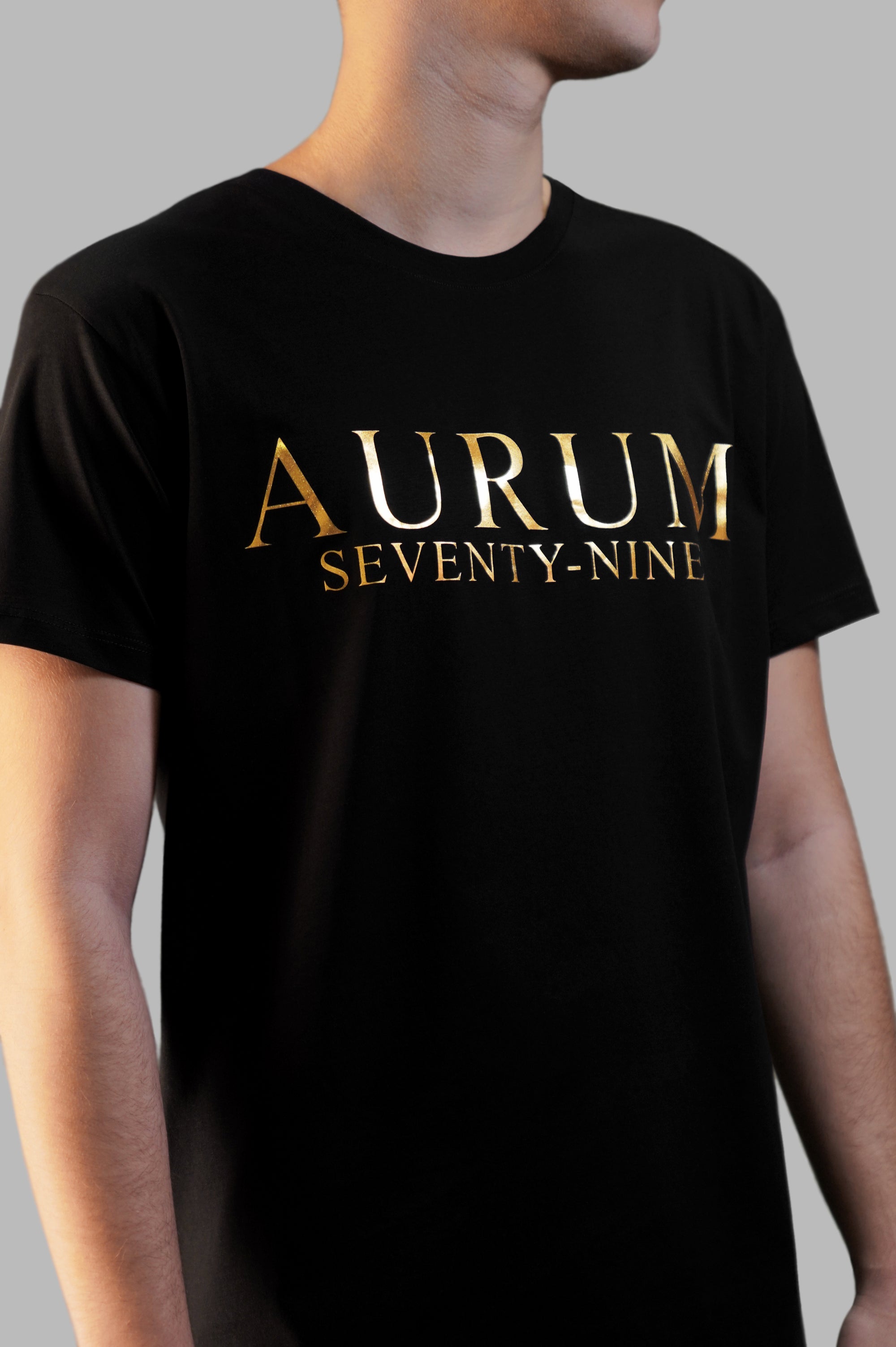 Aurum Seventy-nine: Vista laterale di un modello che indossa la T-shirt Round Neck Aurum Classic Black, con focus sul logo dorato al centro del petto e il girocollo a costina.