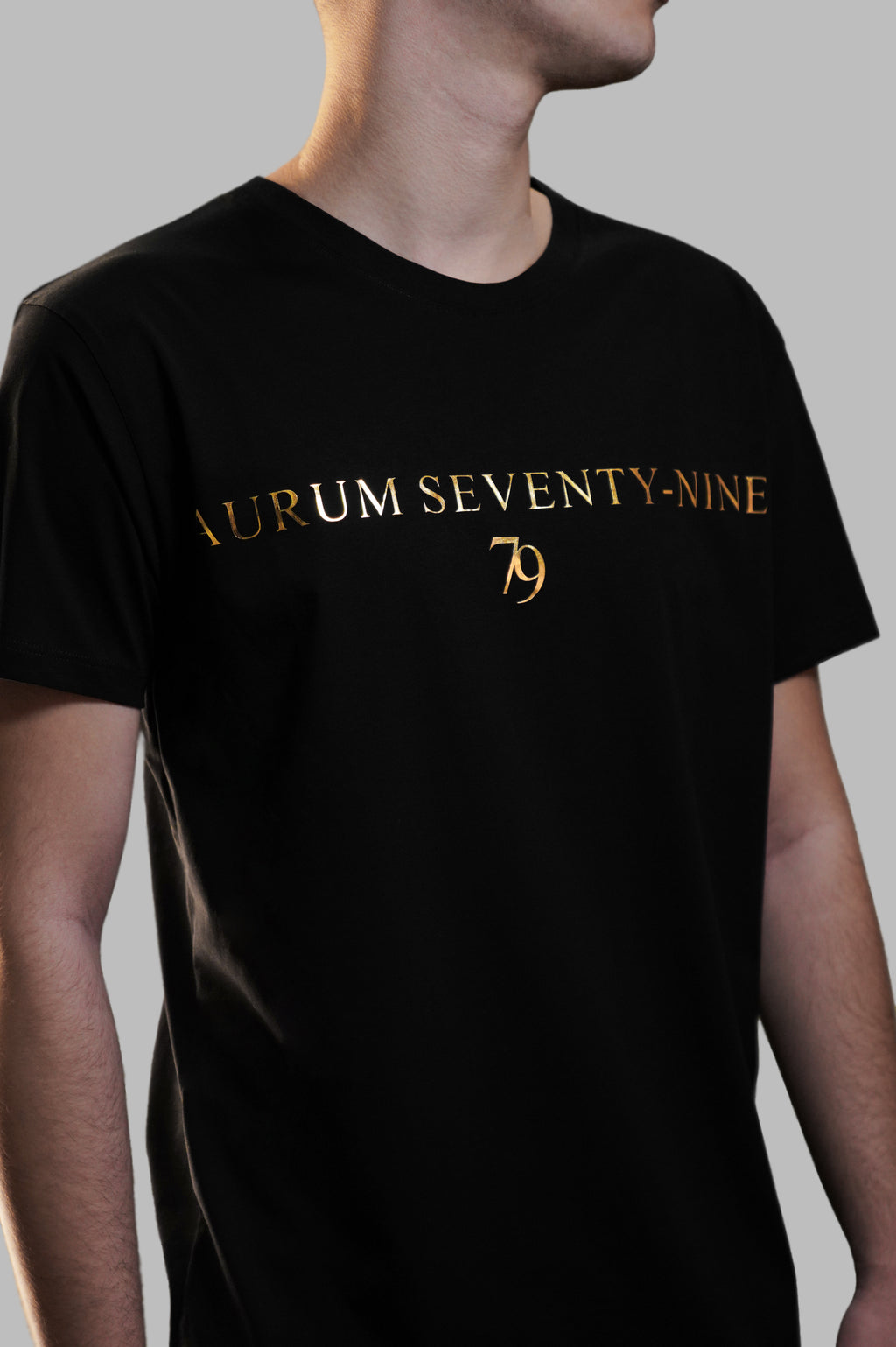 Aurum Seventy-nine: Vista laterale di un modello che indossa la T-shirt Round Neck Aurum Classic 79 Black, con focus sul logo dorato al centro del petto e il girocollo a costina.