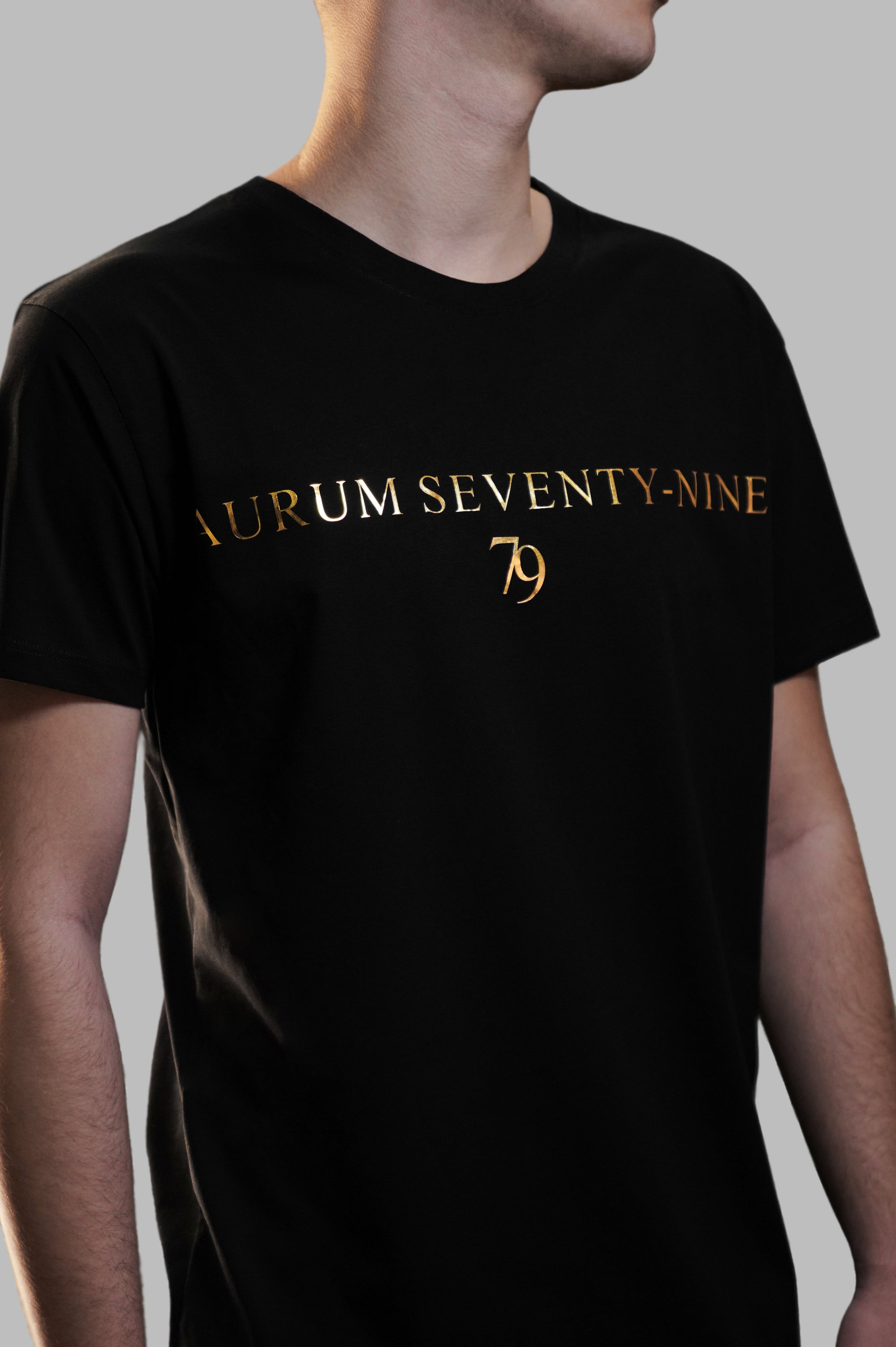 Aurum Seventy-nine: Vista laterale di un modello che indossa la T-shirt Round Neck Aurum Classic 79 Black, con focus sul logo dorato al centro del petto e il girocollo a costina.