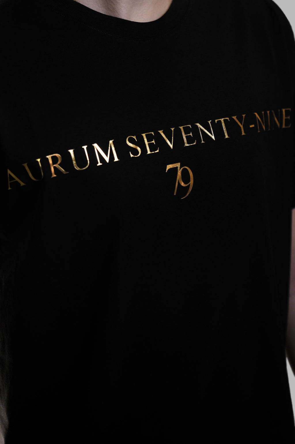Aurum Seventy-nine: Vista leggermente laterale di un modello che indossa la T-shirt Round Neck Aurum Classic 79 Black, con focus sul logo dorato al centro del petto e il girocollo a costina.