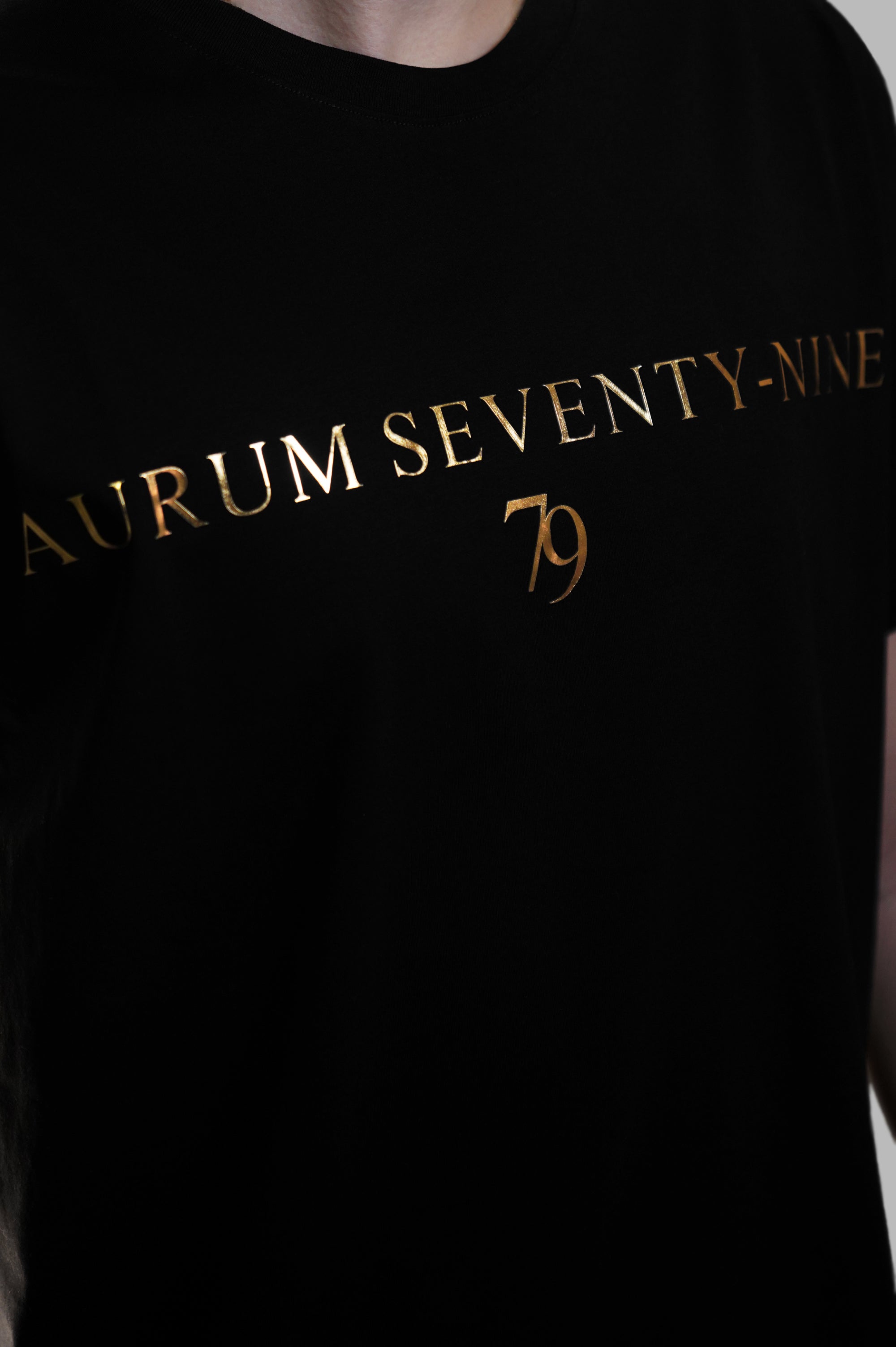 Aurum Seventy-nine: Vista leggermente laterale di un modello che indossa la T-shirt Round Neck Aurum Classic 79 Black, con focus sul logo dorato al centro del petto e il girocollo a costina.
