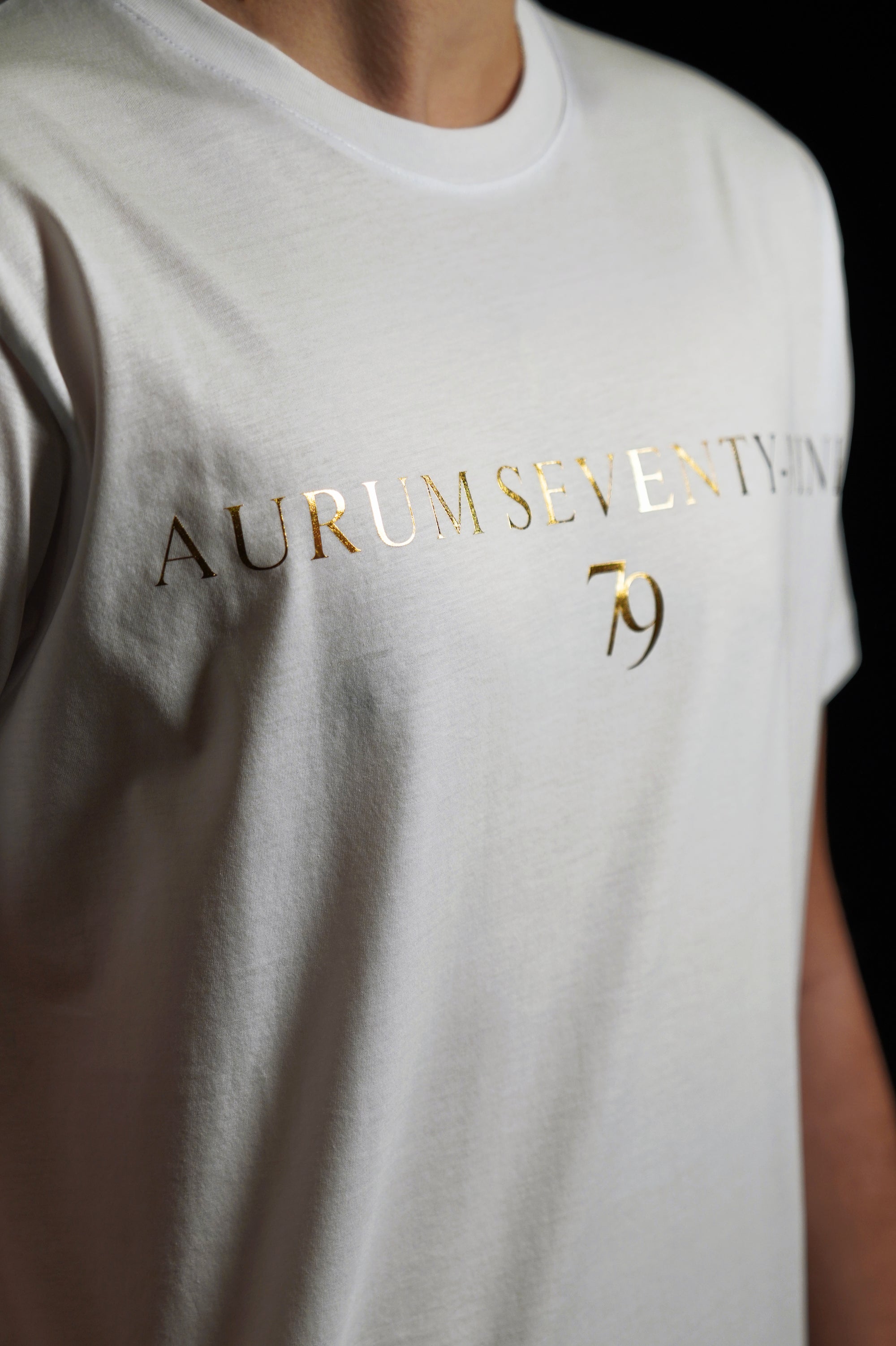 Aurum Seventy-nine: Vista laterale di un modello che indossa la T-shirt Round Neck Aurum Classic 79 White, con focus sul logo dorato al centro del petto e il girocollo a costina.